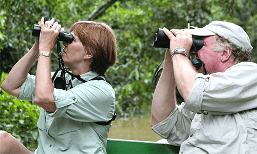 Two visitors peer into binoculars as they sit on a canoe on the Amazon River