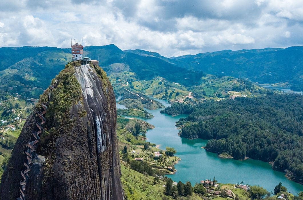 A large rock formation with a staircase built into its side rises above a lush green landscape with a winding river—one of the must-see sights for Colombia tourism, perfect for those seeking unforgettable things to do in Colombia.