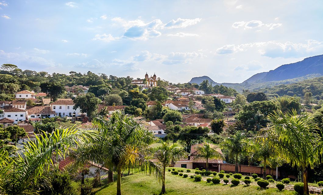 A scenic view of a lush, green town along the historic Route of Gold, with colonial-style white buildings, palm trees, and a church with twin towers in the center—nestled among rolling hills beneath Rio de Janeiro’s partly cloudy sky.