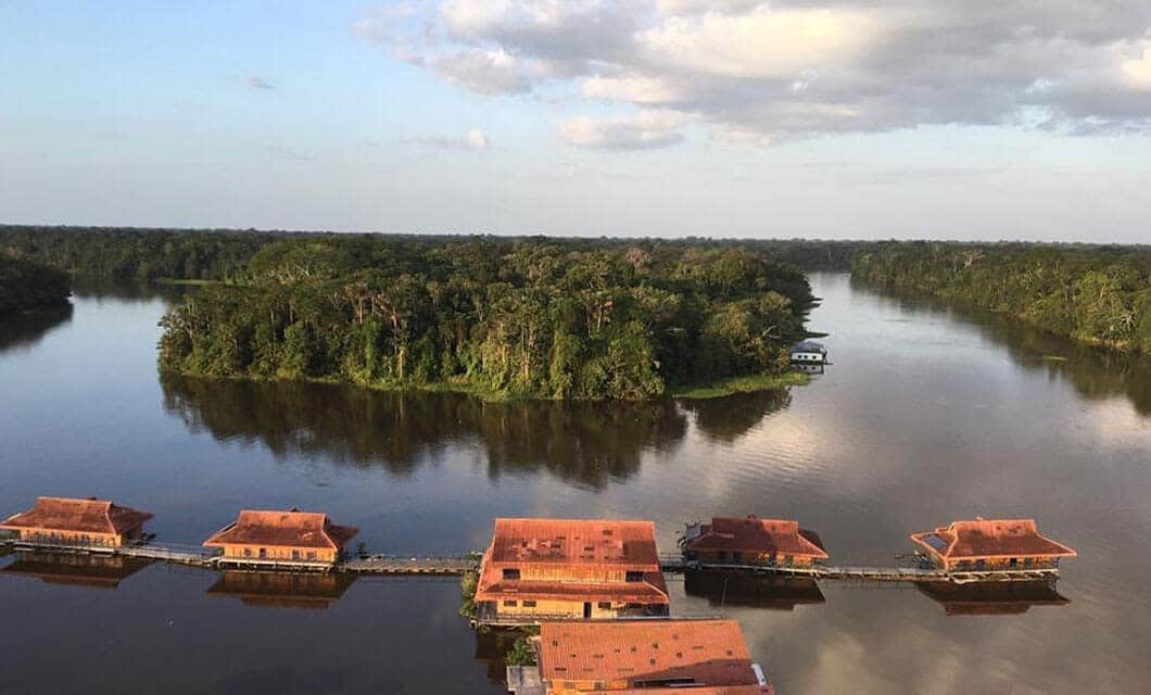 Aerial view of wooden lodges with red roofs built over a calm river, surrounded by dense green forest—perfect for a remote Amazon tour—with an island in the background under a partly cloudy sky.