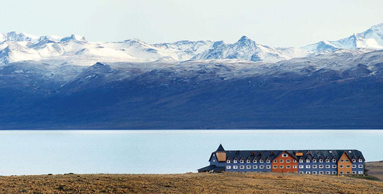 A large lodge-style building sits on grassy plains near a calm lake in Patagonia, with rugged, snow-capped mountains rising in the background under a pale sky—the perfect start to your guided group tour of Patagonia essentials.