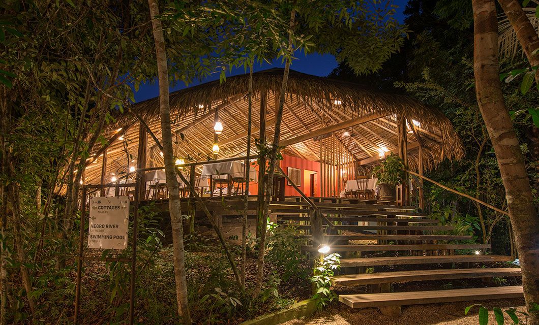 A warmly lit, open-air wooden lodge with a thatched roof is nestled among lush trees at night—an inviting stop on your Brazil Amazon River Cruise—with steps leading up to a dining area and a sign directing guests to the river and swimming pool.