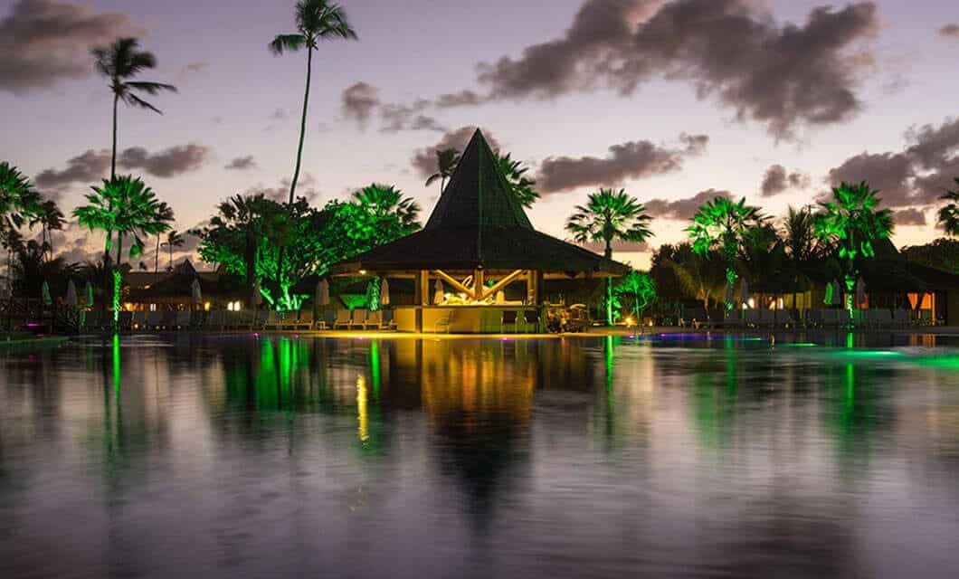 A tropical resort at sunset in Angra dos Reis with palm trees, a thatched-roof pavilion, and green lights reflecting on a calm swimming pool creates a serene and inviting atmosphere at Portobello Hotel.