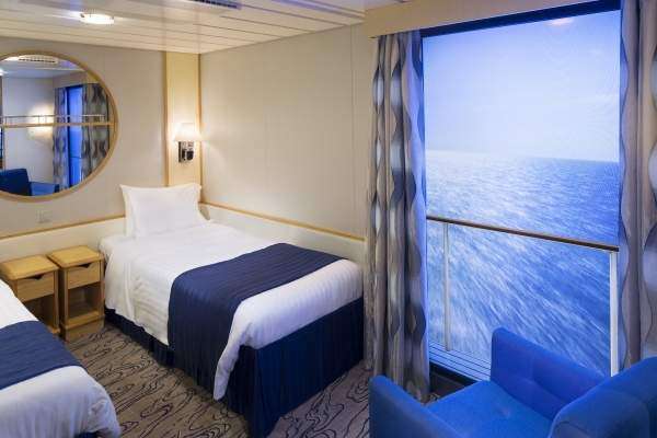 A cruise ship cabin with twin beds dressed in white linens and navy throws, a round mirror on the wall, a blue armchair, and a large window revealing the ocean—perfect comfort for your Galapagos Islands Cruise.