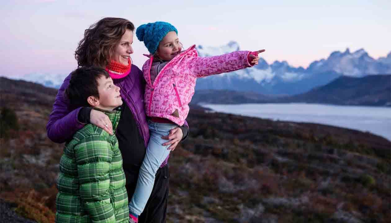A woman hugs two children outdoors near a lake and mountains at sunset. The girl, in a pink jacket and blue hat, points ahead—perhaps toward distant glaciers—as the boy looks too. Their Family Discovery brings them joy.