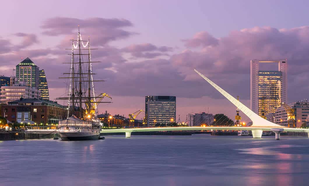 A modern white pedestrian bridge spans a calm river at dusk, with a tall sailing ship docked nearby and city buildings in the background under a purple sky—a striking scene from Wild Argentina.