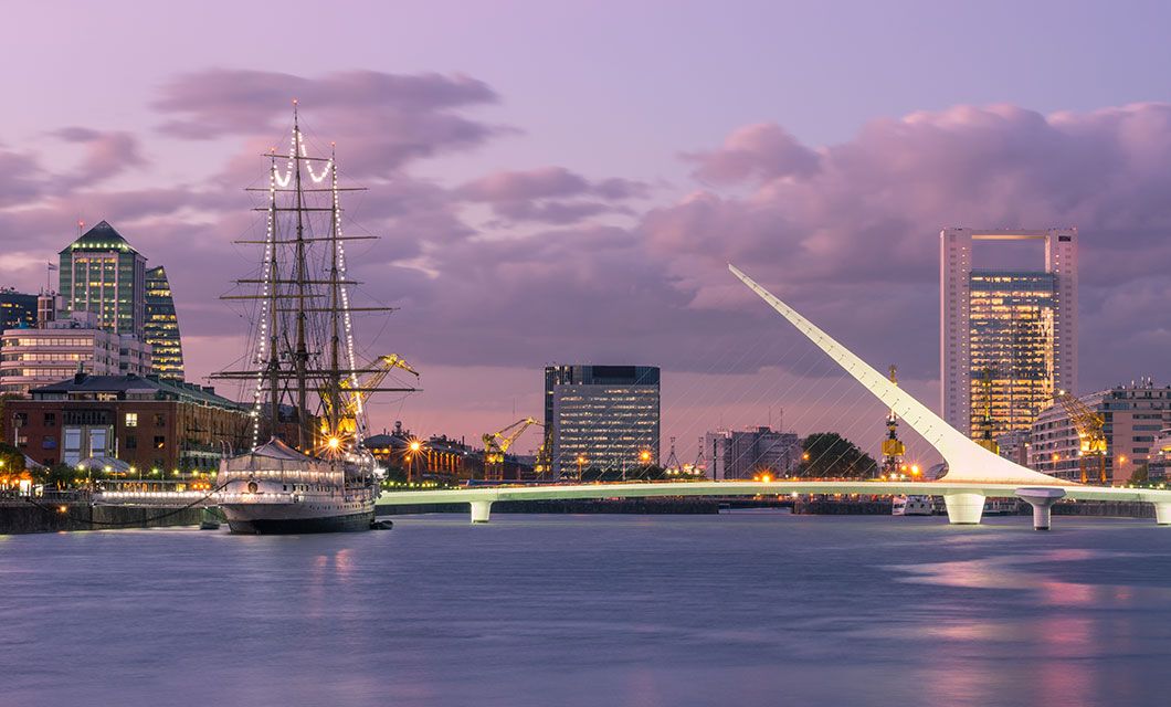A modern white pedestrian bridge spans a calm river at dusk in Buenos Aires, with a historic tall ship docked nearby and city buildings under a pink and purple sky in the background.