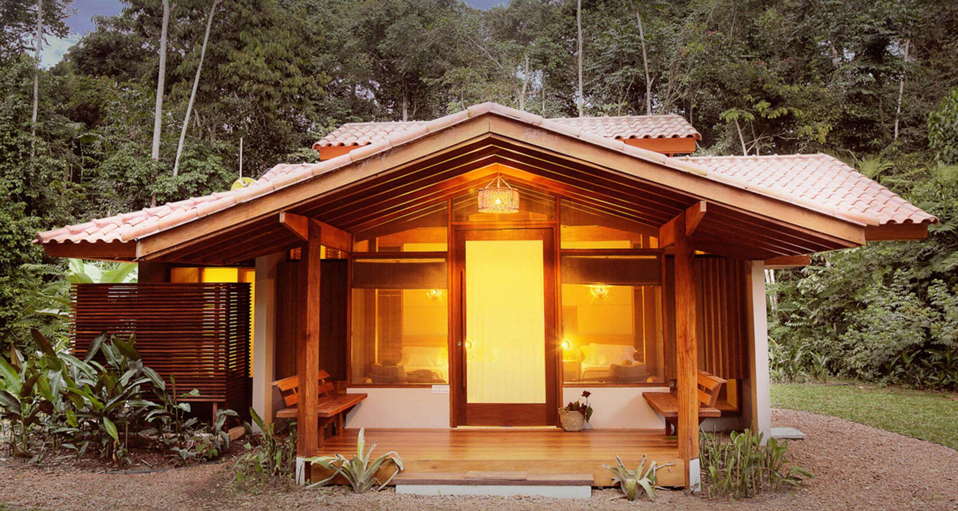 A cozy, single-story wooden house with large windows and a tile roof, warmly lit from within, surrounded by lush green tropical vegetation and forest—perfect for an authentic Amazonian experience.