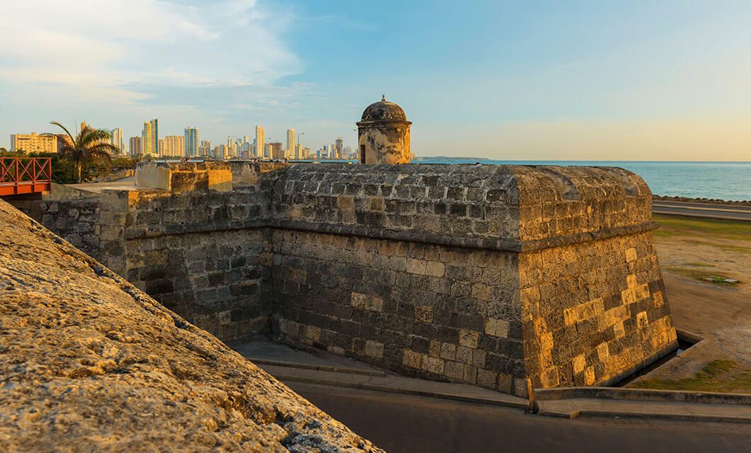 Stone fortification with a rounded watchtower overlooks the sea in Colombia, with a modern city skyline visible in the background under a clear sky at sunset—a perfect start before your botanical tour.