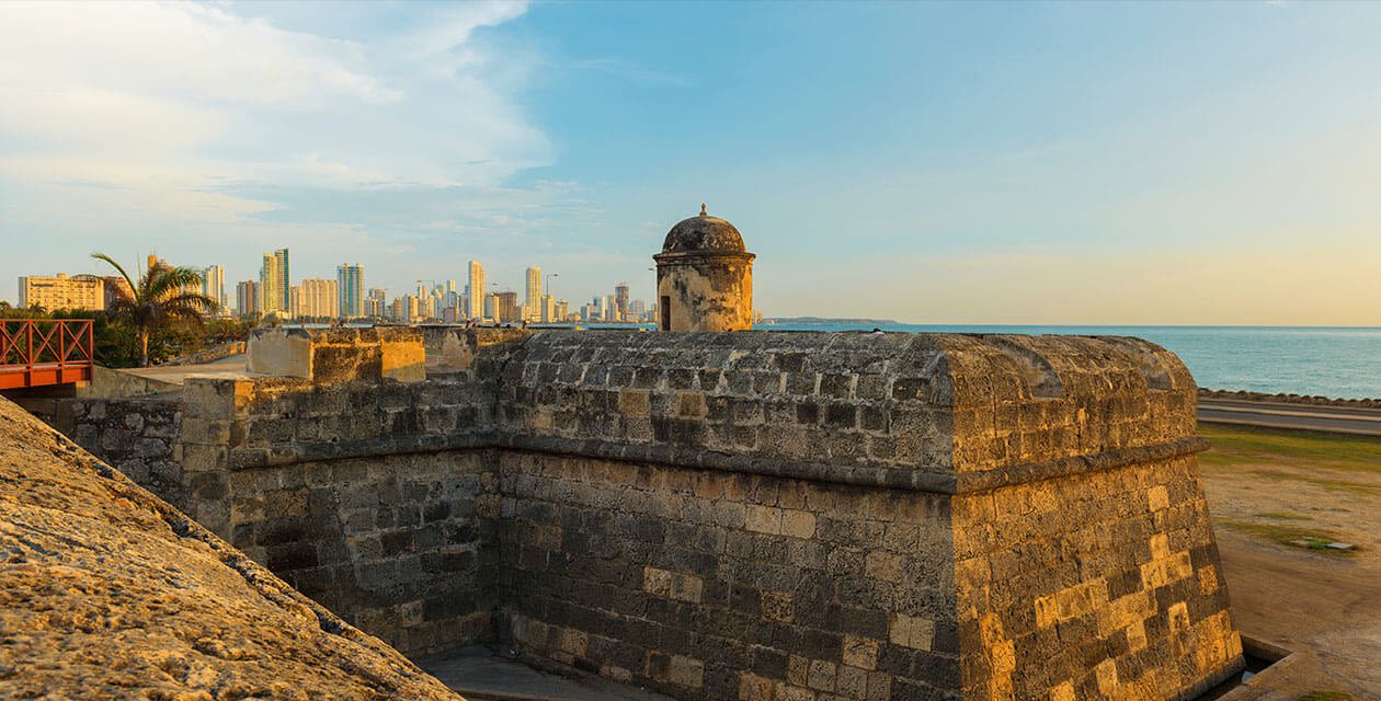 Stone fortification with a guard tower overlooks the sea, while Cartagena's modern skyline and palm trees rise in the background under a blue sky—a perfect start to your Cartagena tour or Black History Tour.