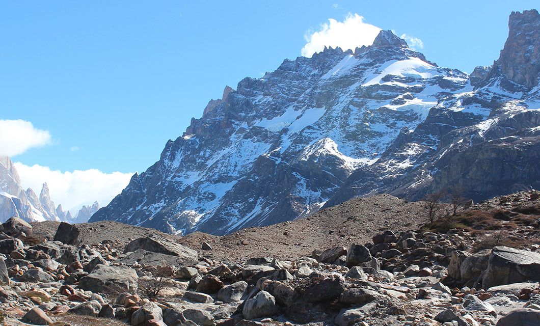 Snow-capped mountain with rugged, rocky slopes under a bright blue sky in Argentina, seen from a foreground of scattered rocks and sparse vegetation—perfect for True Explorers seeking breathtaking scenery.