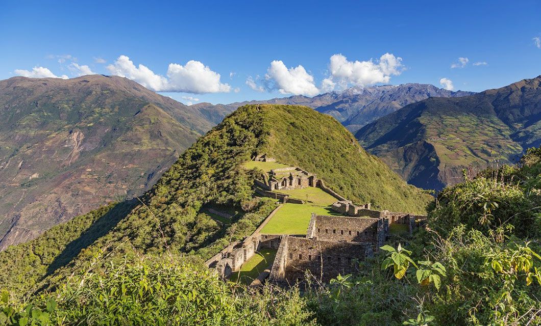 Ancient stone ruins sit on a lush green mountain ridge along the Choquequirao trek, under a bright blue sky, surrounded by distant peaks and valleys covered in vegetation.