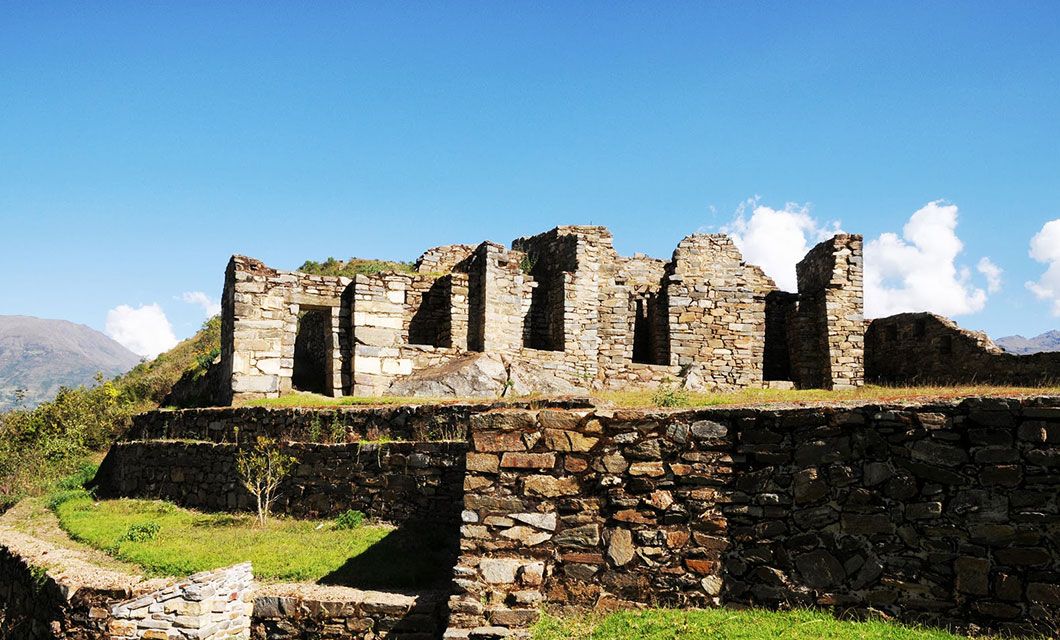 Ancient stone ruins of a building sit atop a grassy terrace, surrounded by green hills under a bright blue sky—just one stunning scene along the Choquequirao trek in Peru.
