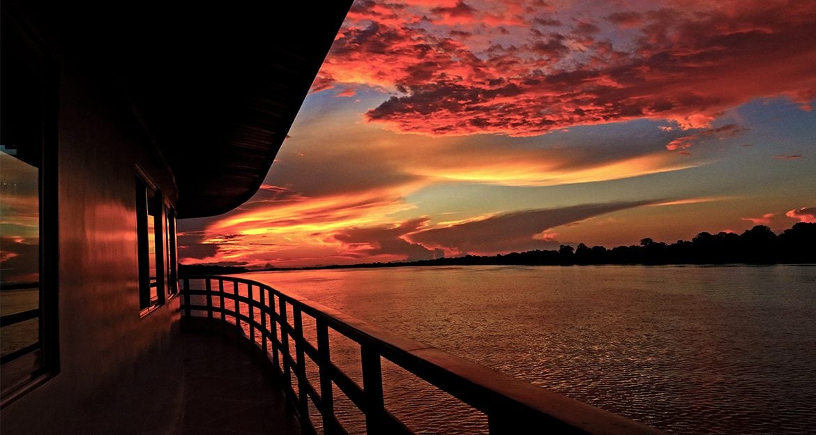 A vivid sunset over the calm Amazon River, viewed from an Amazon Clipper Cruise with curved railings and windows reflecting dramatic orange, red, and purple clouds. Trees line the distant shore, completing this unforgettable Amazon Cruises experience.