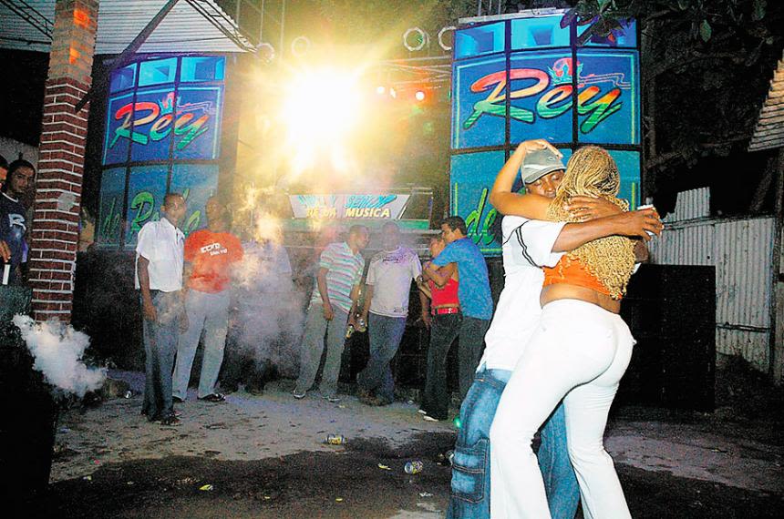 People dance closely in the foreground at an outdoor party or club in Cartagena, while others stand and watch near a stage with colorful lights and signs reading Rey. Smoke and bright lighting fill the lively scene on this festive tour.