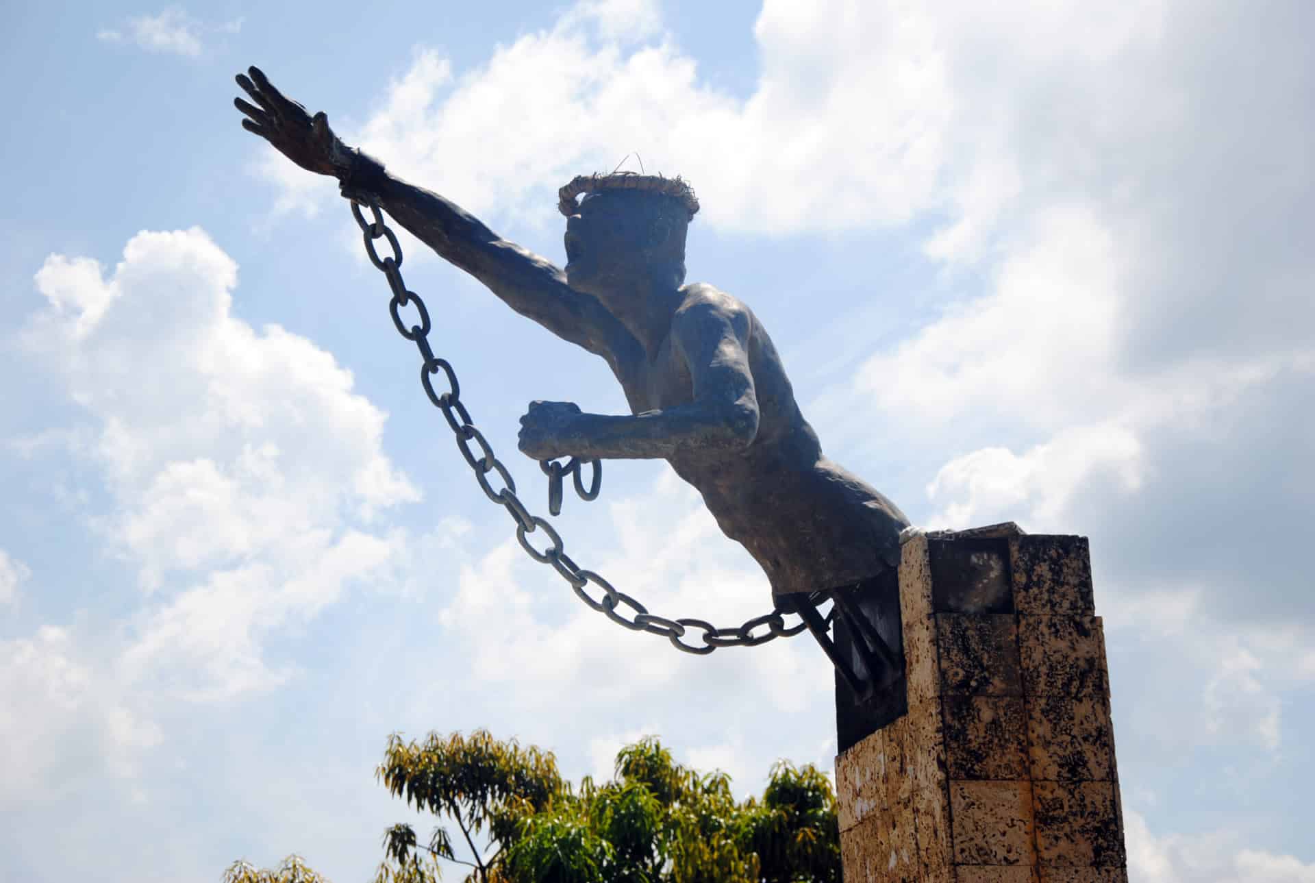 A bronze statue of a man with a broken chain on his wrist reaches upward, symbolizing freedom from slavery. Featured on the Black History Tour in Cartagena, it stands on a stone pedestal against a backdrop of sky and clouds.
