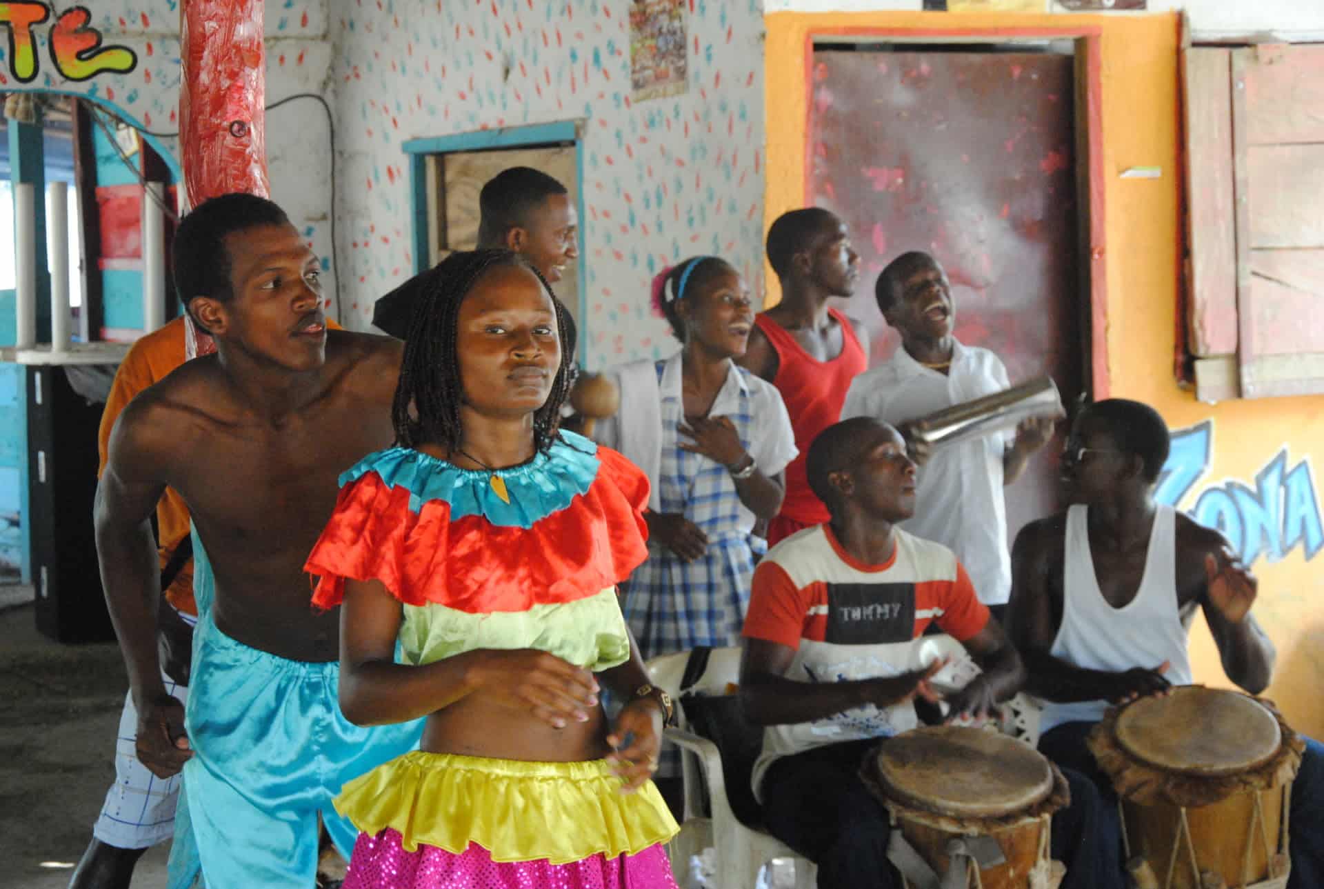 A group of people, some playing drums and others singing or dancing, gather in a colorful indoor setting in Cartagena. Two dancers in vibrant costumes lead the lively scene, part of an engaging Black History Tour celebrating rich cultural traditions.