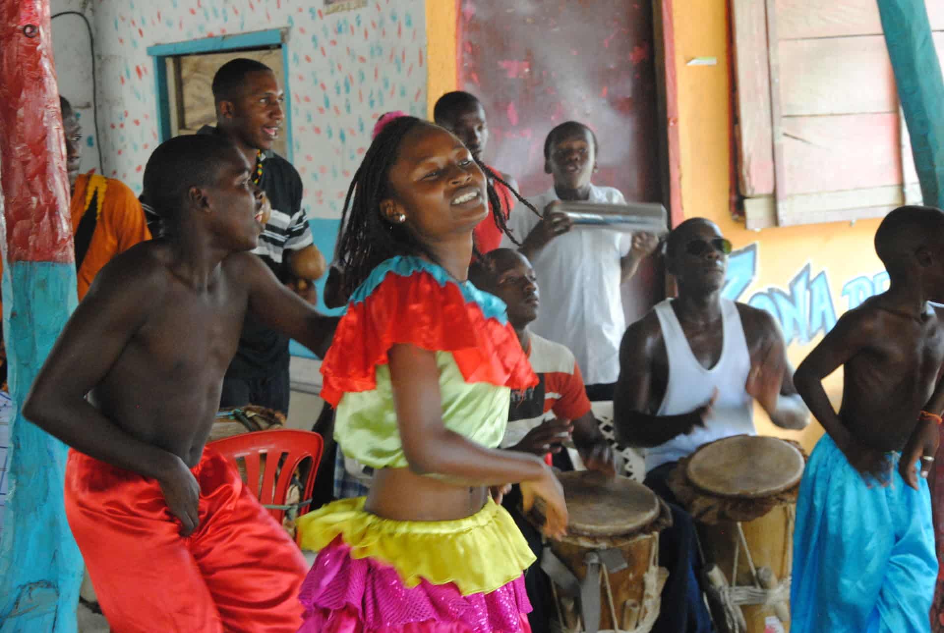 A group of people joyfully dance and play drums indoors in Cartagena. A young girl in a colorful ruffled outfit smiles at the center, surrounded by boys and men in casual clothes, capturing the spirit of a Black History Tour celebration.