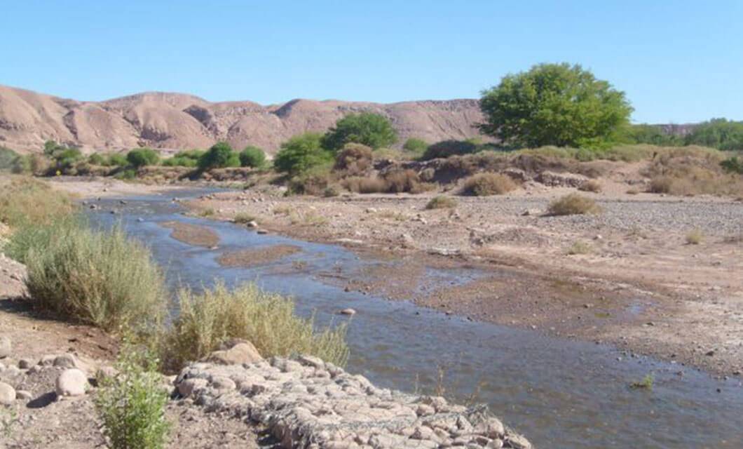 A small, shallow stream flows through a dry, rocky landscape with sparse green bushes and trees. In the background, brown hills rise beneath a clear blue sky—hinting at a quiet journey across the boundaries of time.