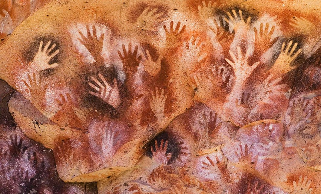 A cave wall along the Carretera Austral, covered with numerous hand stencils in shades of brown, red, and white, overlapping each other to create a dense, textured pattern—an iconic sight on a Patagonia tour.