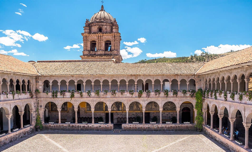 A historic courtyard in Cusco with arched walkways and a central open area, surrounded by a two-story stone building. In the background, a large domed bell tower rises against a bright blue sky with scattered clouds.