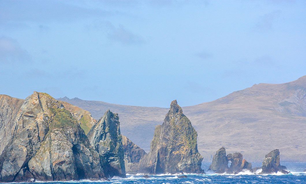 Rocky sea stacks rise from the ocean near a rugged coastline, with waves crashing at their base—an iconic scene reminiscent of a Patagonia cruise departing from Ushuaia, set against distant hills and a partly cloudy blue sky.