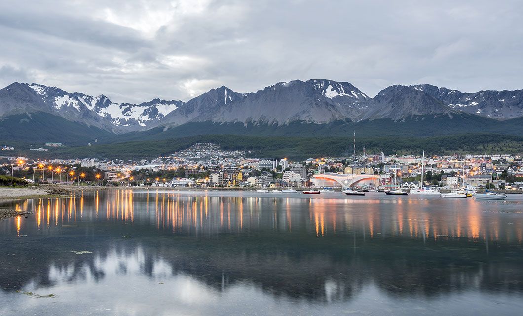 A calm waterfront city in Ushuaia with snow-capped mountains in the background. Buildings and streetlights reflect on the still water under a cloudy sky, creating a serene and picturesque scene perfect for a Patagonia cruise adventure.