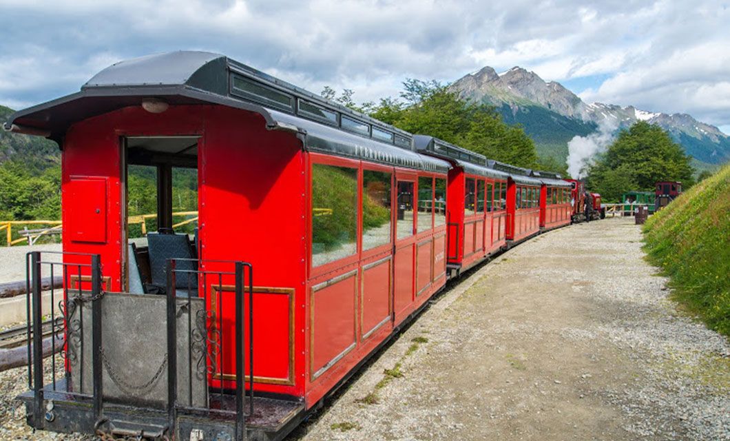 A bright red passenger train travels along a track in a scenic, mountainous area with green trees and snow-capped peaks in the background, recalling the breathtaking landscapes near Torres del Paine under a partly cloudy sky.