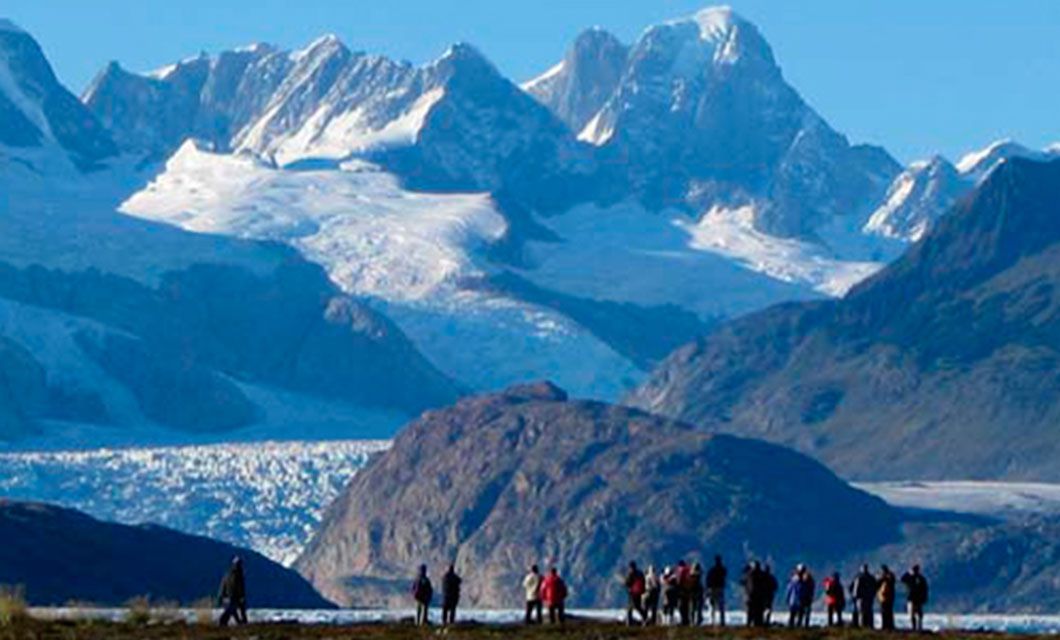 A group of people stands on a hill in the foreground, looking toward snow-covered mountains and a glacier under a clear blue sky, capturing the breathtaking beauty of Torres del Paine.