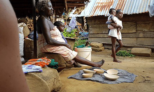 An elderly woman sits outside with a young child on her lap, surrounded by bowls on a mat. Nearby, a girl holds another child beside rustic wooden and corrugated metal buildings—a glimpse into daily life on a Three Guianas adventure tour.