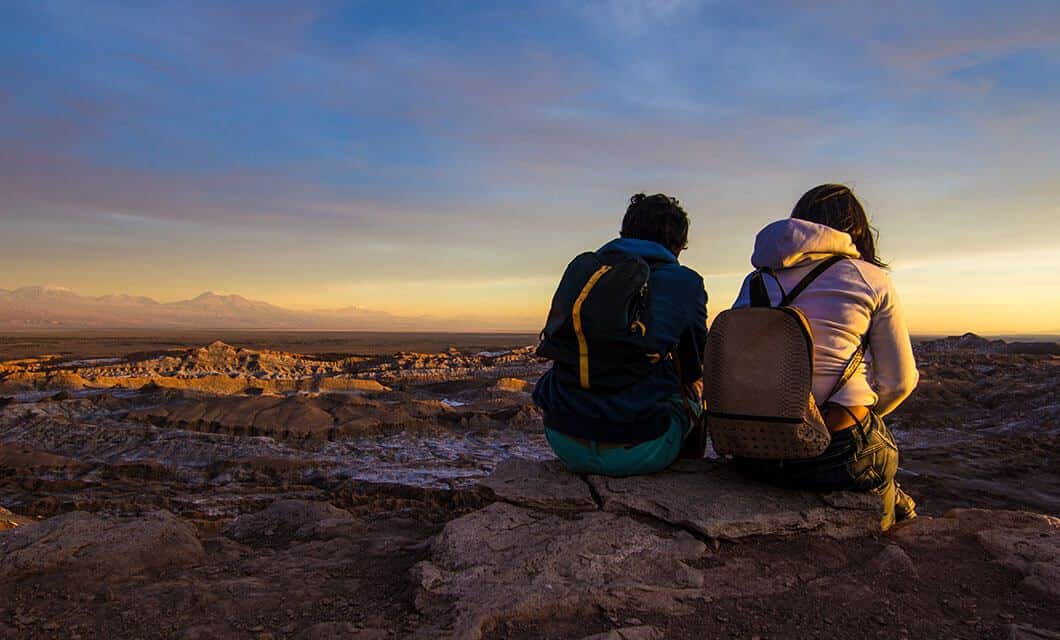 Two people with backpacks sit on a rocky ledge, pausing mid-journey to watch the sunset over a vast desert. Distant mountains and a colorful sky evoke the Boundaries of Time.