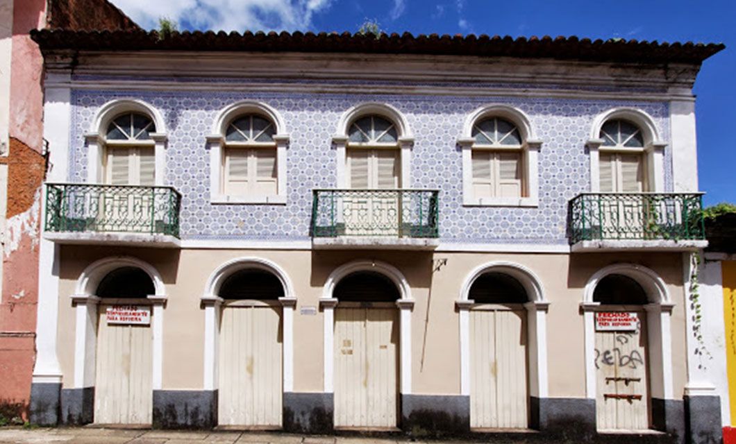 Two-story building with blue-and-white patterned tiles on the upper facade, arched windows and doors, white shutters, two small balconies with green railings—ideal for a luxury adventure stay or as a base for your Lencois Maranhenses tour. For sale signs on boarded lower doors.