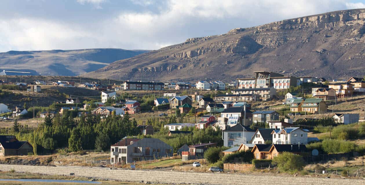 A small town with colorful houses and buildings scattered on a hillside, surrounded by trees and shrubs, sits beneath rocky mountains under a partly cloudy sky—the perfect setting for a Family Journey at the End of The World.