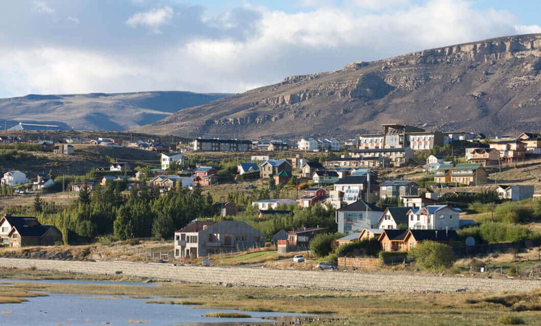 A small town with scattered houses and greenery sits at the base of rocky, barren hills under a partly cloudy sky. In Patagonia, a river or lake along the foreground reflects the landscape—perfect for a group tour or guided tour experience.