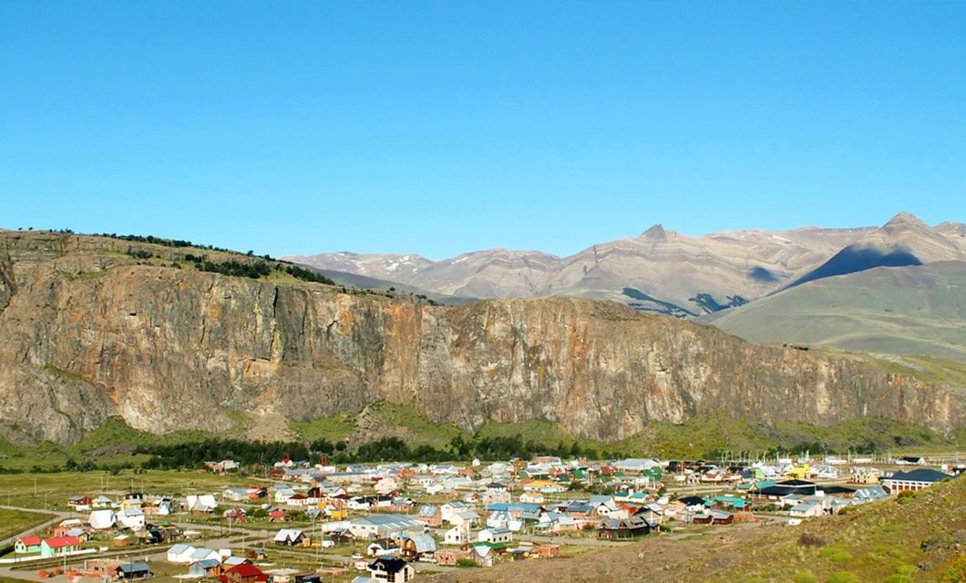 A small town with colorful rooftops sits at the base of tall rocky cliffs, surrounded by green fields and distant mountains—a perfect scene for a Patagonia guided group tour under a clear blue sky.