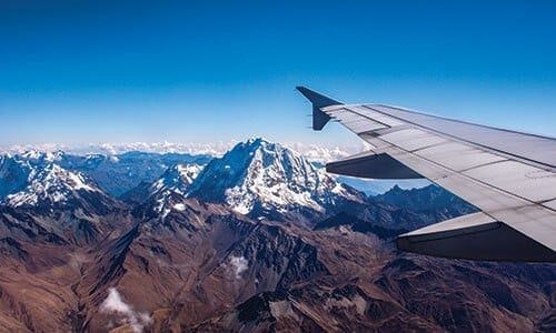 A view from an airplane window shows a wing over snow-capped mountains and rugged terrain under a clear blue sky, capturing the breathtaking beauty of South American Wonders—an unforgettable scene for any honeymoon South America adventure.