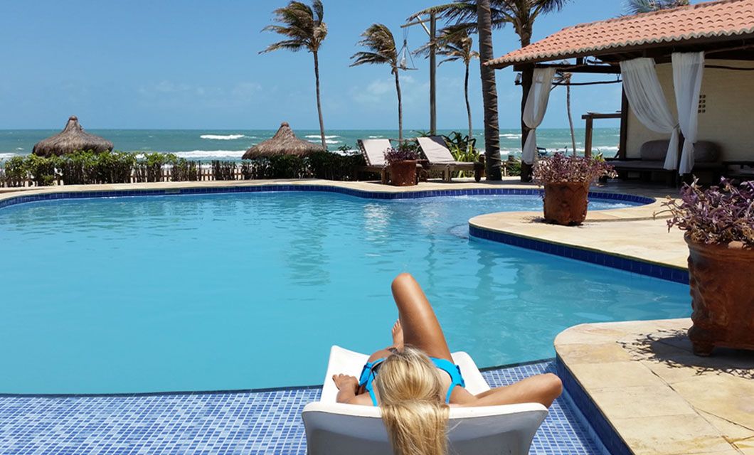 A woman in a blue bikini lounges on a chair by a pool, facing the ocean. Palm trees sway in the breeze, with cabanas and beach chairs beyond—perfect for a luxury adventure tour under the clear, sunny sky.