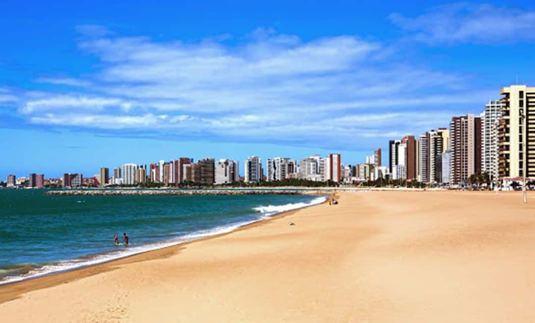 A wide sandy beach with gentle waves, a few people near the shoreline, and a city skyline of tall modern buildings under a blue sky with scattered clouds—perfect for a luxury adventure tour.