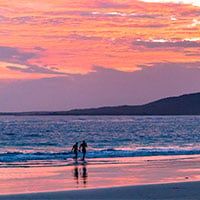 Two people walk along the shore of a beach at sunset, with vibrant pink and orange clouds in the sky and gentle waves in the background—reminiscent of tranquil evenings on a Manatee Explorer Amazon Cruise.