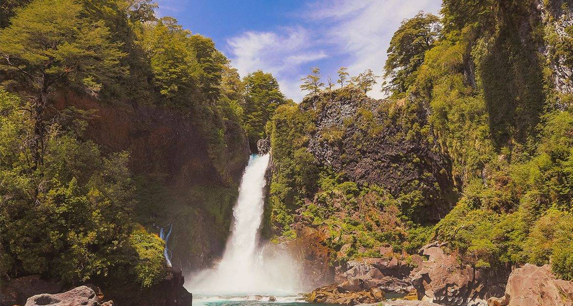 A tall waterfall cascades down a rocky cliff in the Huilo-Huilo Reserve, Chile, surrounded by lush green trees and dense vegetation, with water splashing into a turquoise pool below under a bright sky.