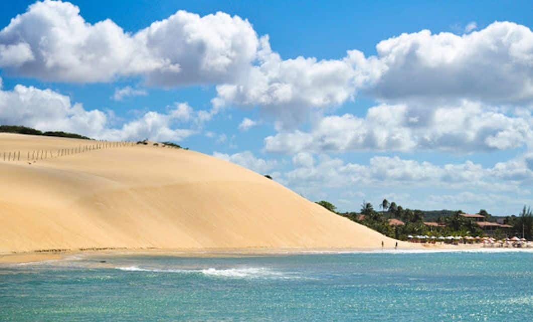 A large golden sand dune at Lencois Maranhenses meets a blue ocean under a partly cloudy sky, with gentle waves along the shore—an inviting scene perfect for a luxury adventure tour amid distant buildings and palm trees.