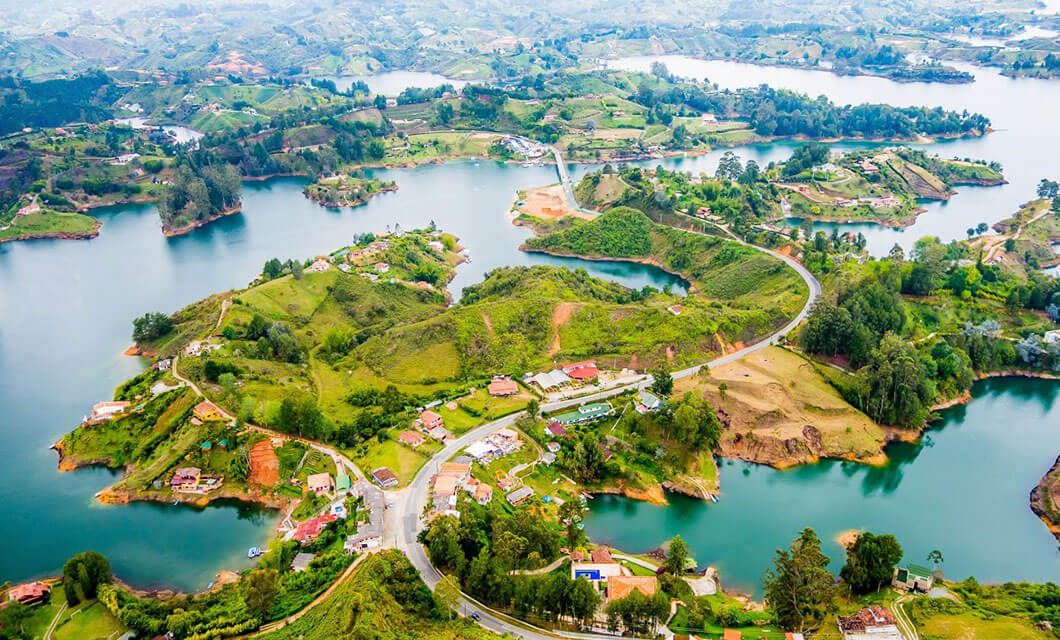 Aerial view of a lush, green landscape in Colombia with winding roads, scattered houses, and blue water lakes weaving between peninsulas and hills—an inviting scene perfect for a Botanical Tour under a clear sky.