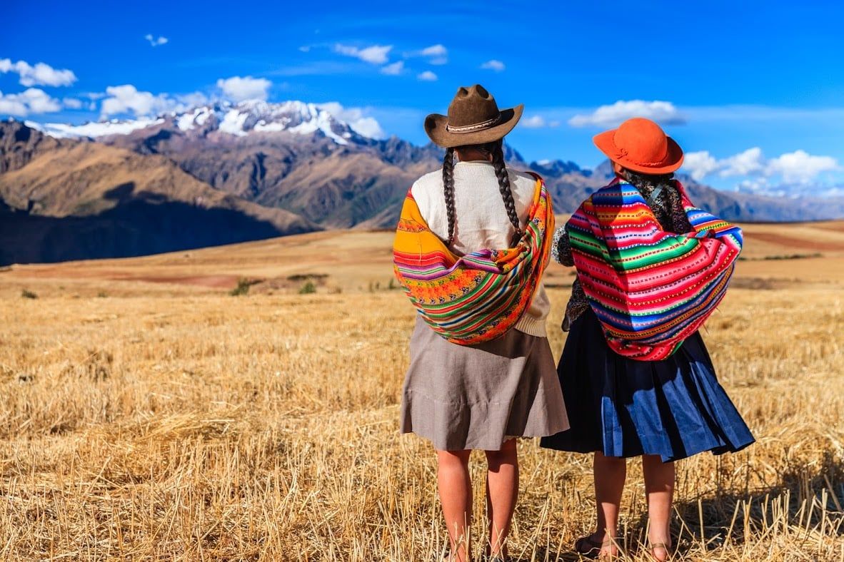 Two women in traditional Andean clothing and hats stand in a golden field, gazing at snow-capped mountains under a bright blue sky—a perfect scene capturing the magic of South American Wonders.