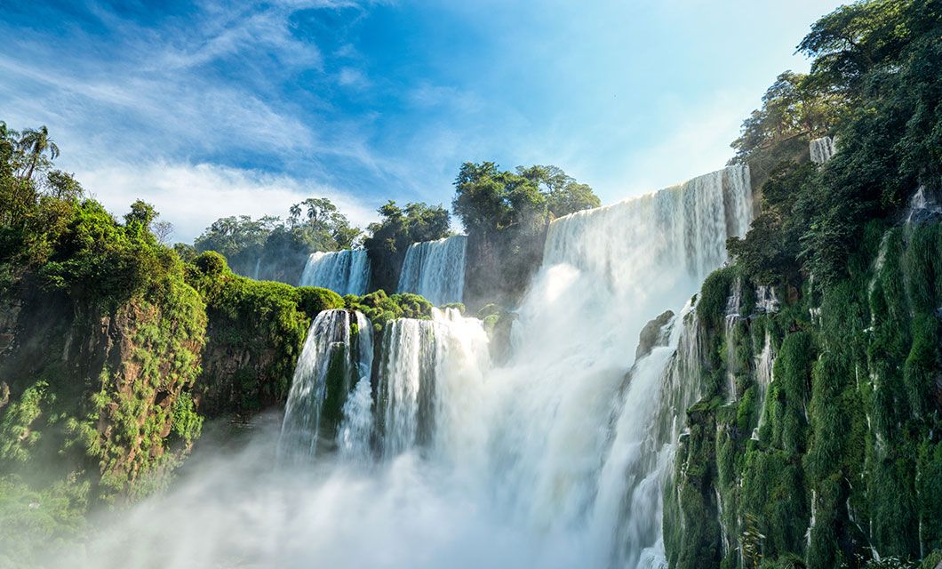 A powerful waterfall cascades over lush, green cliffs under a bright blue sky—an unforgettable highlight of any Brazil tour, surrounded by dense tropical forest and mist rising from the falling water.