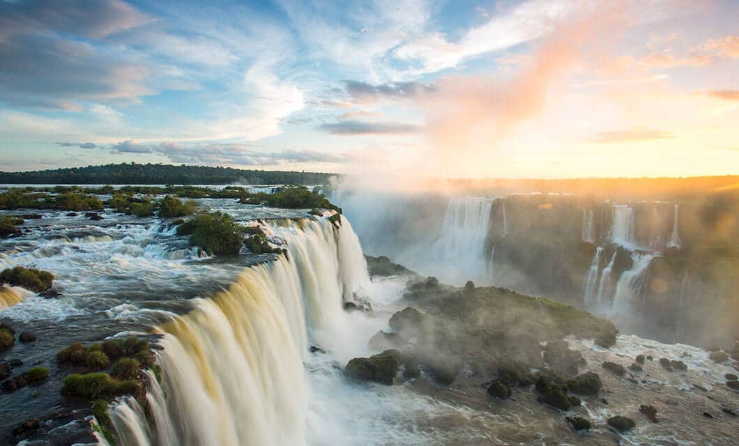 A wide view of powerful waterfalls cascading over cliffs with mist rising, surrounded by lush greenery in wild Argentina, under a partly cloudy sky at sunrise or sunset.