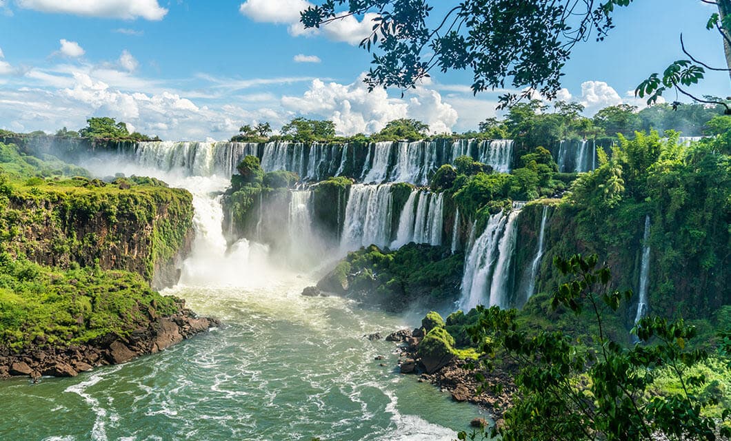 A wide view of Iguazu Falls in Wild Argentina shows multiple waterfalls cascading over lush green cliffs into a river, with mist rising and trees framing the scene under a partly cloudy blue sky.