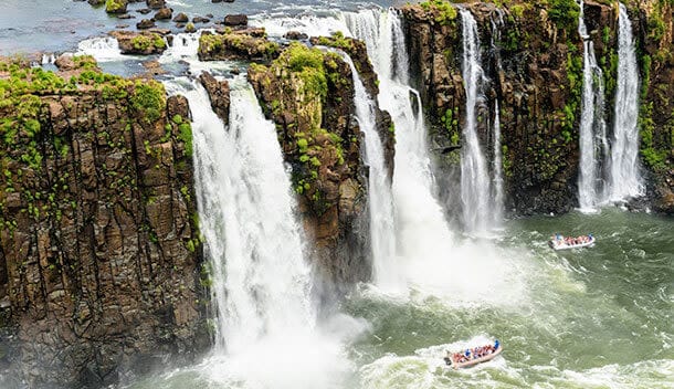 Several powerful waterfalls cascade over rocky cliffs into a river below, with three tour boats filled with people approaching the falls, surrounded by lush greenery and scenic lakes.