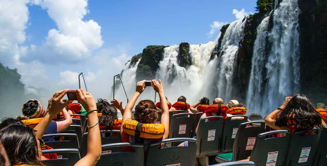 True Explorers in Argentina wear orange life jackets as they sit on a boat, capturing photos and enjoying a close view of large, cascading waterfalls surrounded by lush greenery under a bright blue sky with scattered clouds.