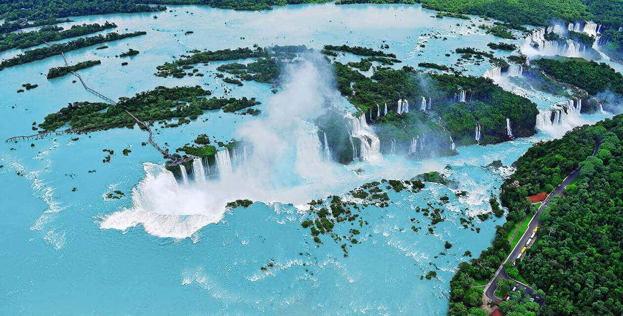 Aerial view of Iguazu Falls, one of South America's natural wonders, with multiple waterfalls cascading into turquoise water, surrounded by lush green forests and small islands. Mist rises from the powerful falls.