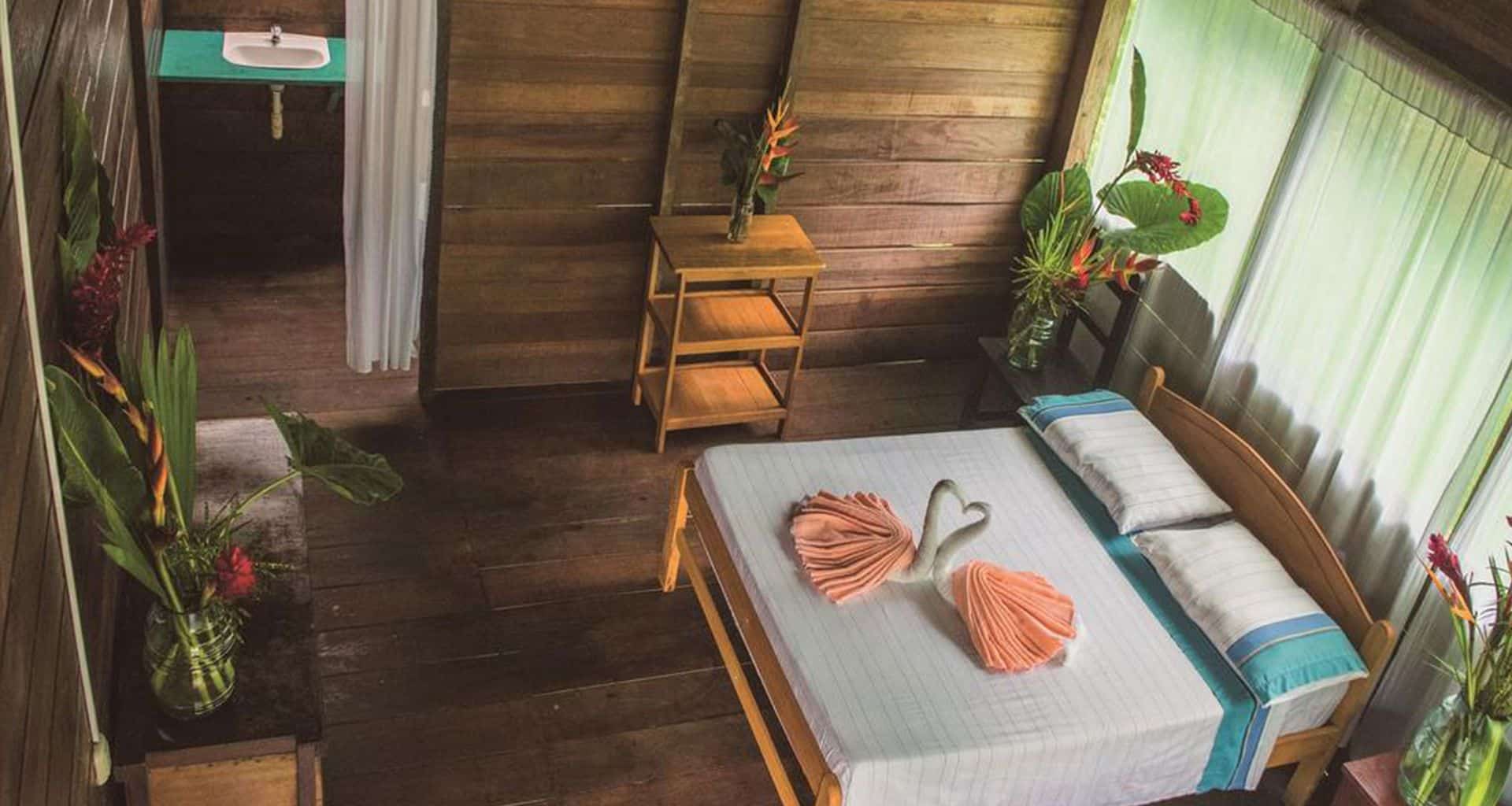 A cozy wooden room at Inotawa Lodge with a double bed, pastel linens, and towels folded into swan shapes. Bright green plants and flowers add charm, while large curtained windows frame this inviting Tambopata Amazon lodge retreat.